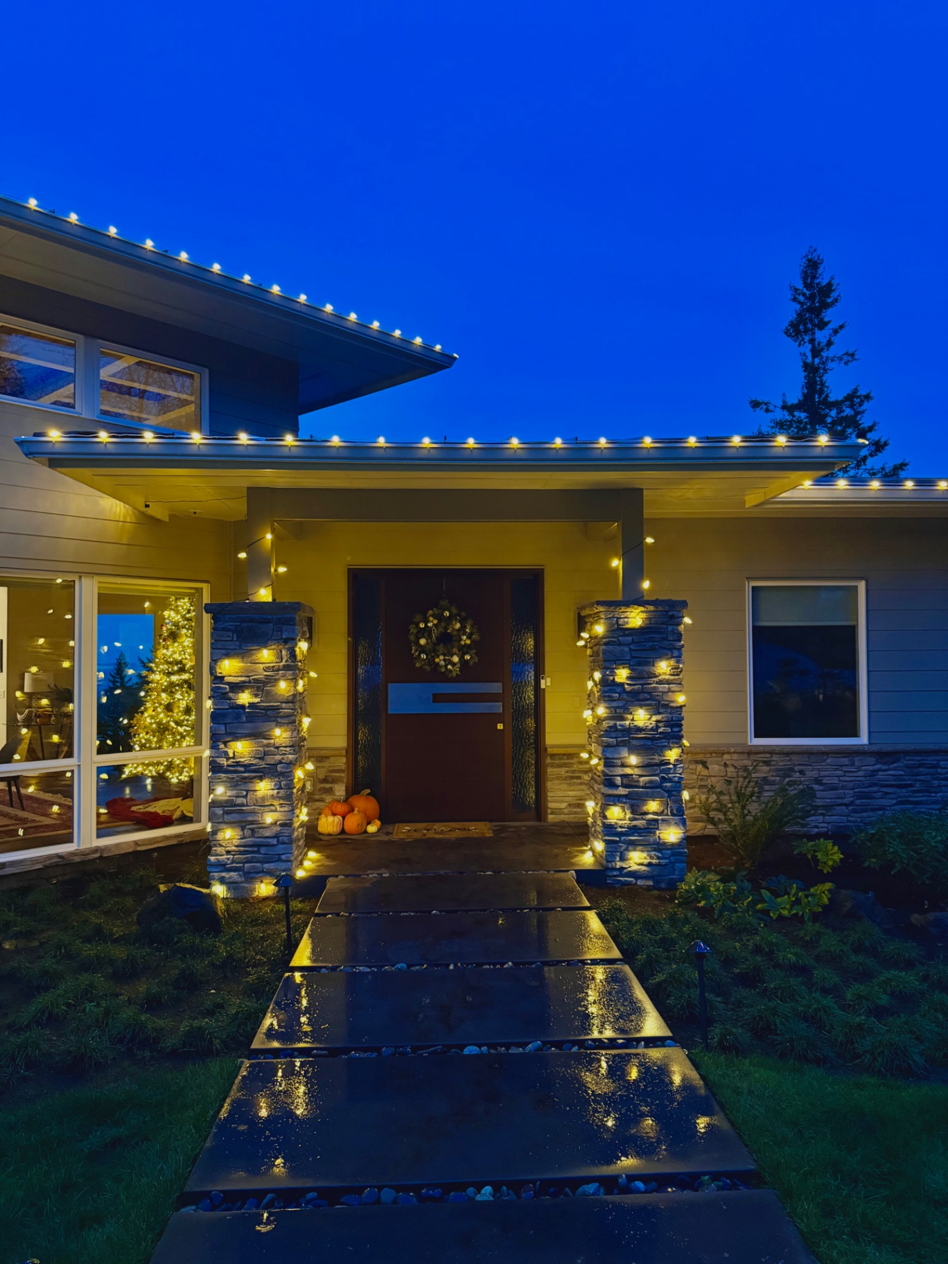 Warm-white Christmas lights wrapping stone entry columns and the roofline of a modern Oregon home at dusk