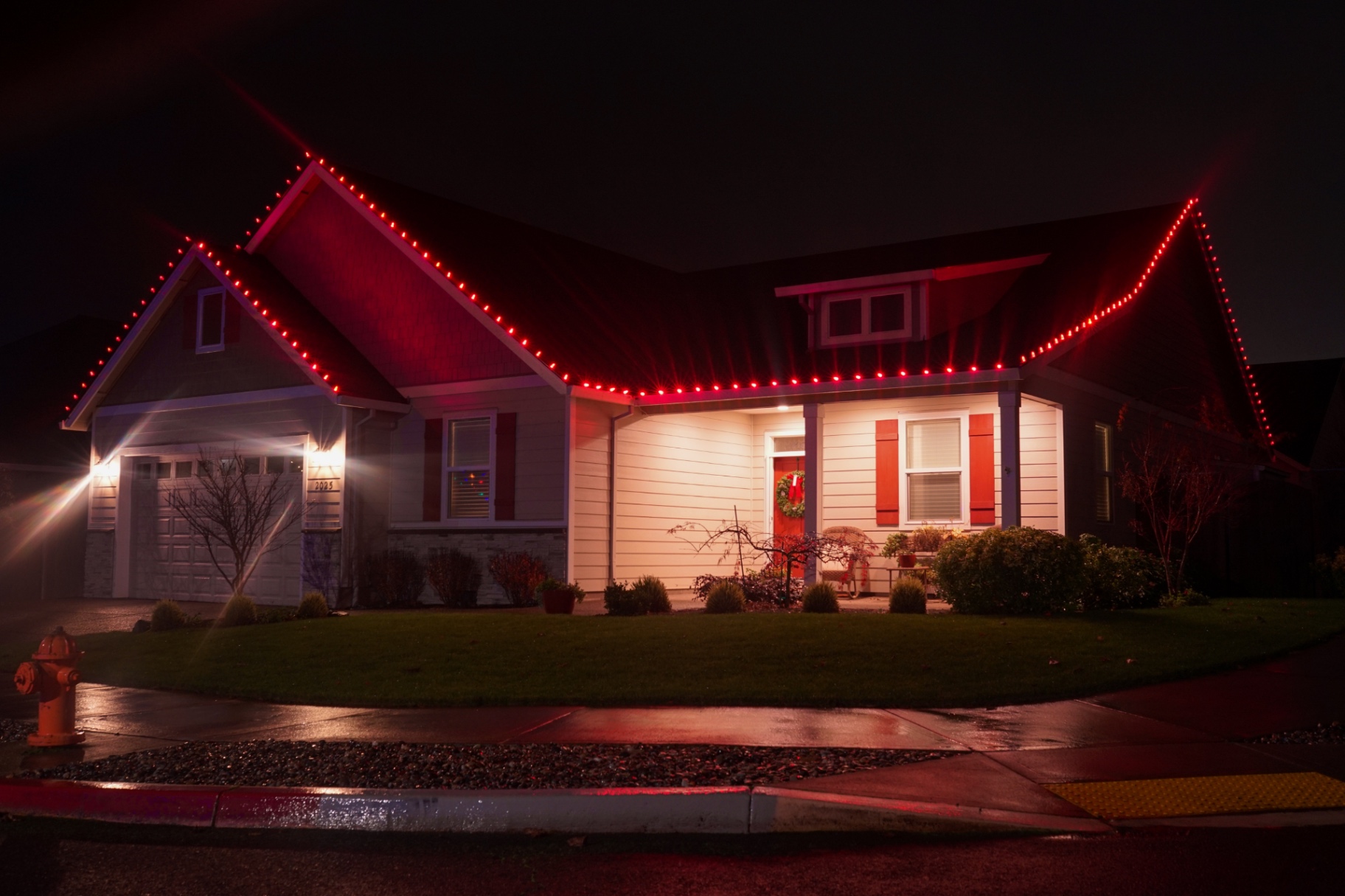 Craftsman home outlined in solid red C9 Christmas lights reflecting on a wet street at night