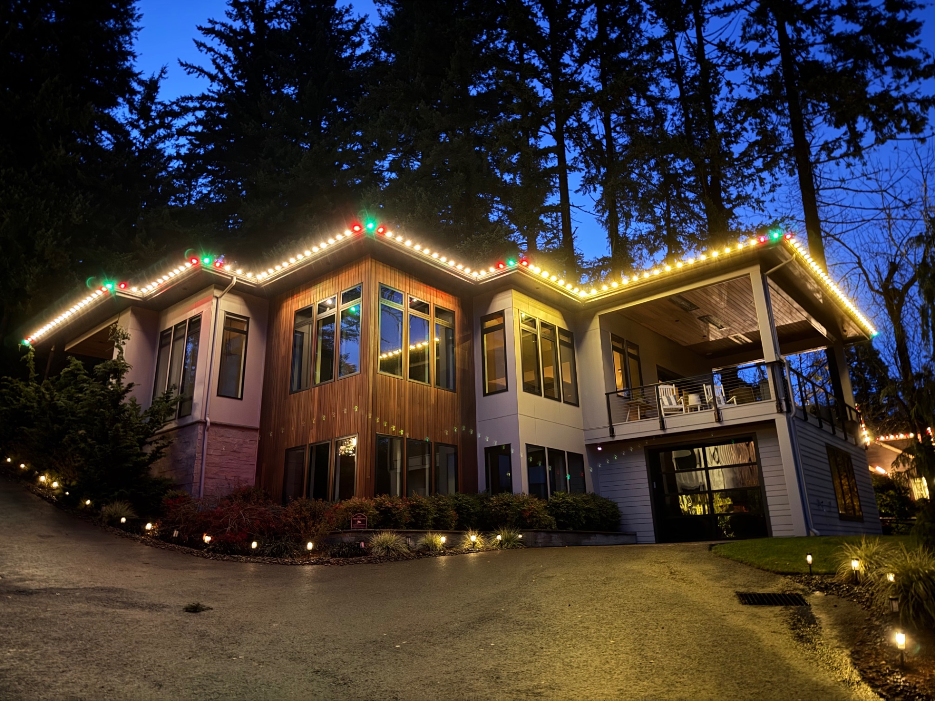 Cedar and modern hillside home outlined with warm-white C9 Christmas lights and red/green peak accents at dusk